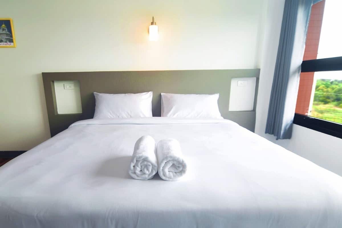 A hotel room with a neatly made bed, two pillows, a wooden chair, and a view through a large window.