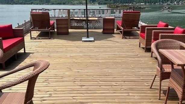 A wooden deck with wicker furniture and a white umbrella, overlooking a lake and forested hills. - The Mughal Palace Eco Luxury Houseboat  (Srinagar)