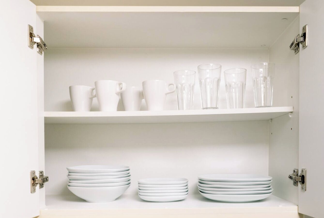 A white cabinet with two shelves, each holding stacks of white plates and clear glassware.