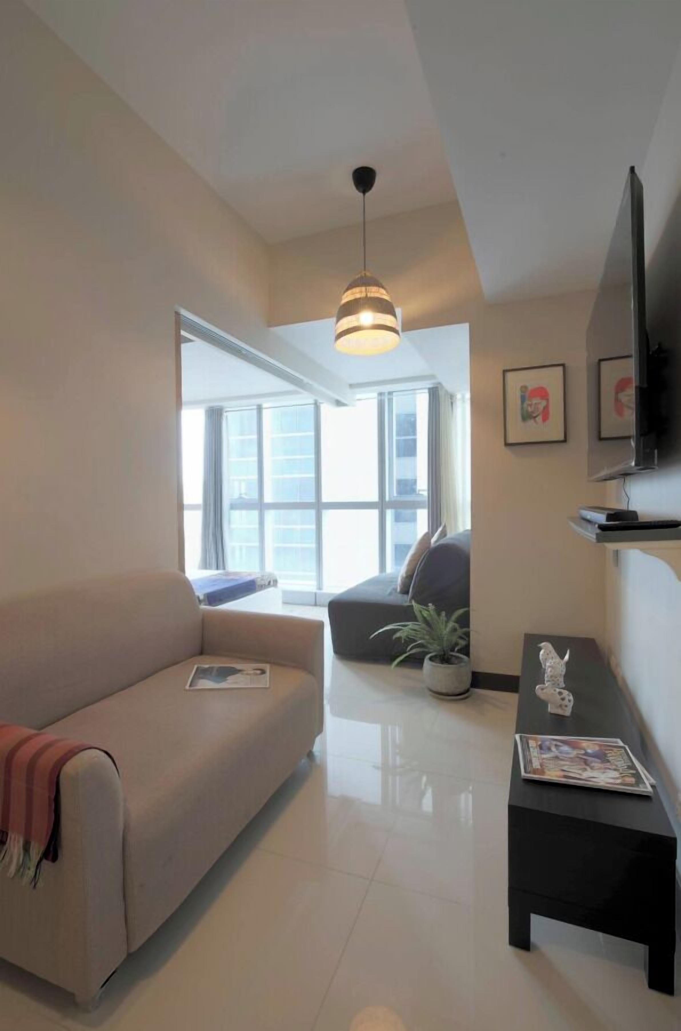 Uma sala de estar moderna com um sofá, uma mesa de centro e uma televisão. Há uma janela grande que oferece luz natural e vista para o exterior.