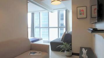 Uma sala de estar moderna com um sofá, uma mesa de centro e uma televisão. Há uma janela grande que oferece luz natural e vista para o exterior.