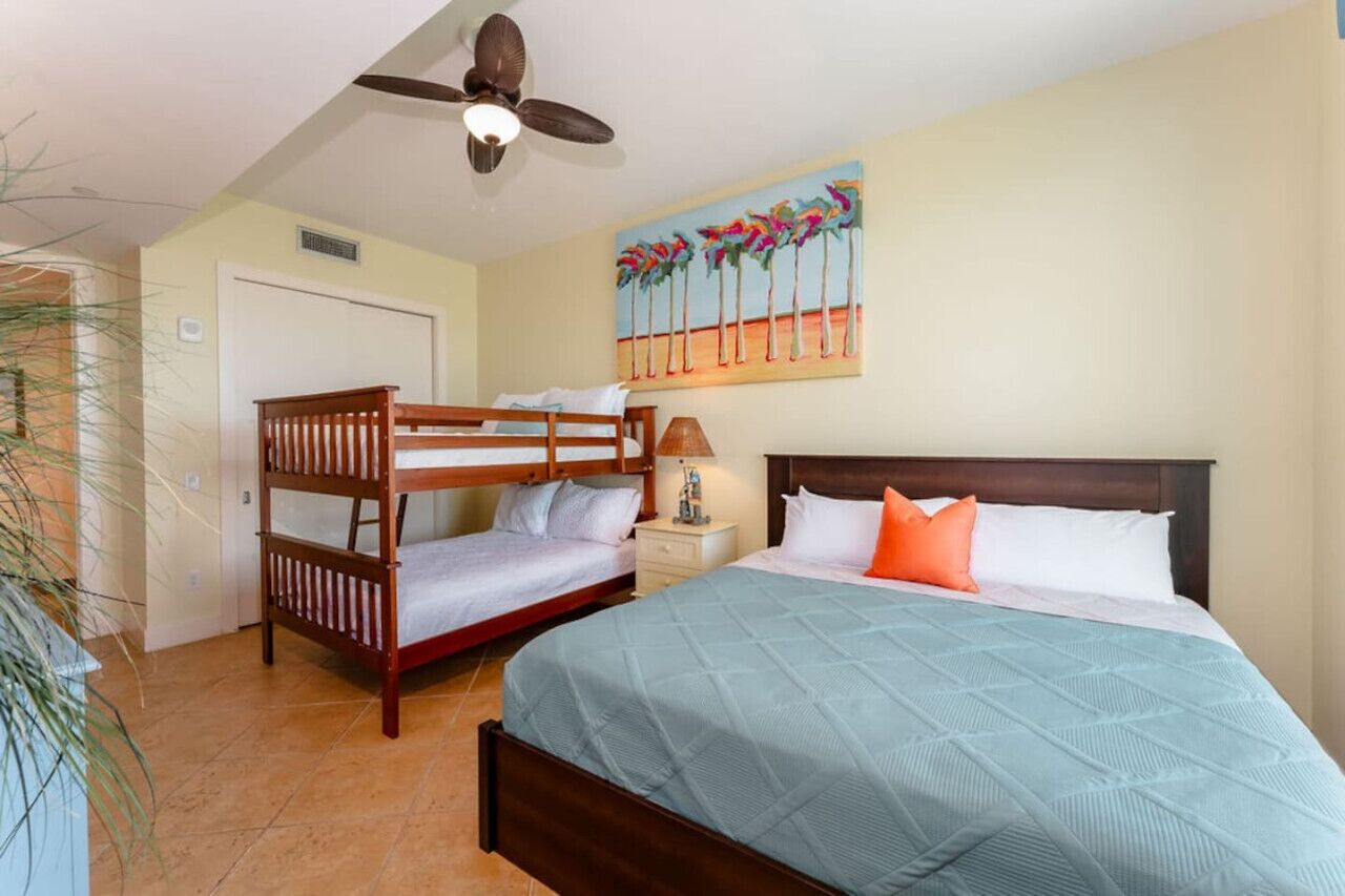 Una habitación de hotel con literas, una cama individual, un ventilador de techo, un cuadro de palmeras y una lámpara de noche.