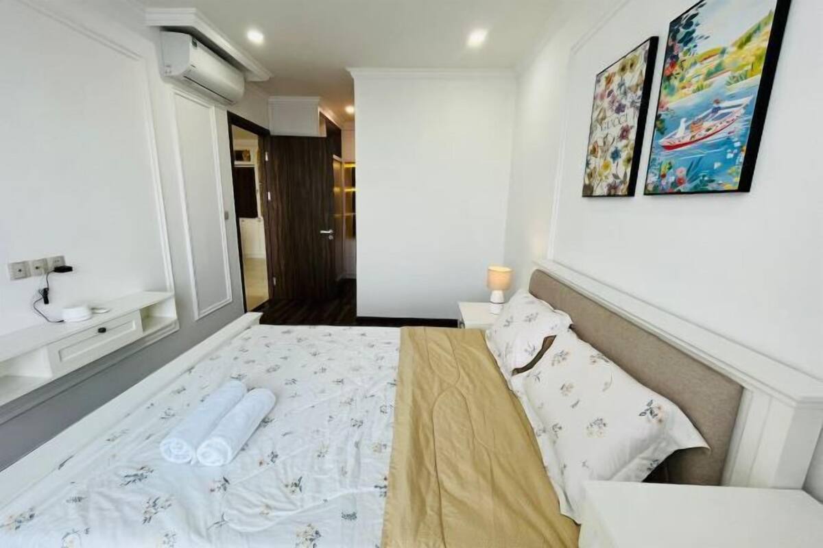 Habitación de hotel con cama, mesita de noche, aire acondicionado de pared, lámpara y dos cuadros enmarcados.