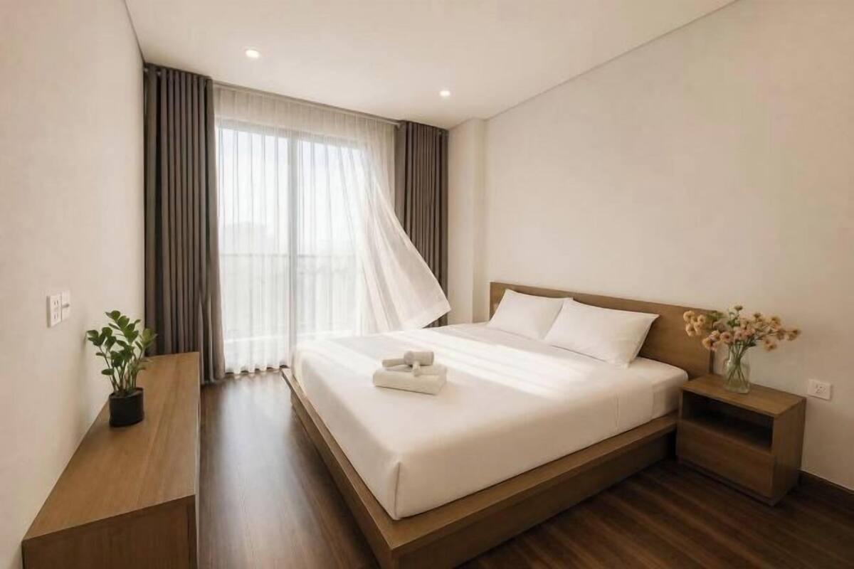 Una habitación de hotel con una cama grande, una mesita de noche, cortinas en la ventana y una planta pequeña.