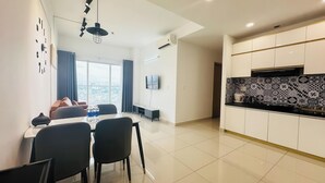A modern living room with a sofa, a dining table with chairs, a TV mounted on the wall, and a large window with curtains. - Charmington La Pointe - Central Suites D (Ho Chi Minh City)