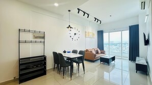 A modern living room with a dining area, a sofa, a coffee table, a TV, and a large window with a city view. - Charmington La Pointe - Central Suites D (Ho Chi Minh City)