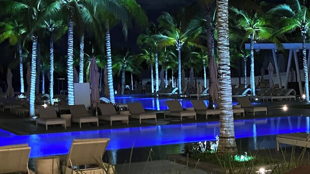 Zwembadgedeelte van een resort bij nacht, met palmbomen en ligstoelen.