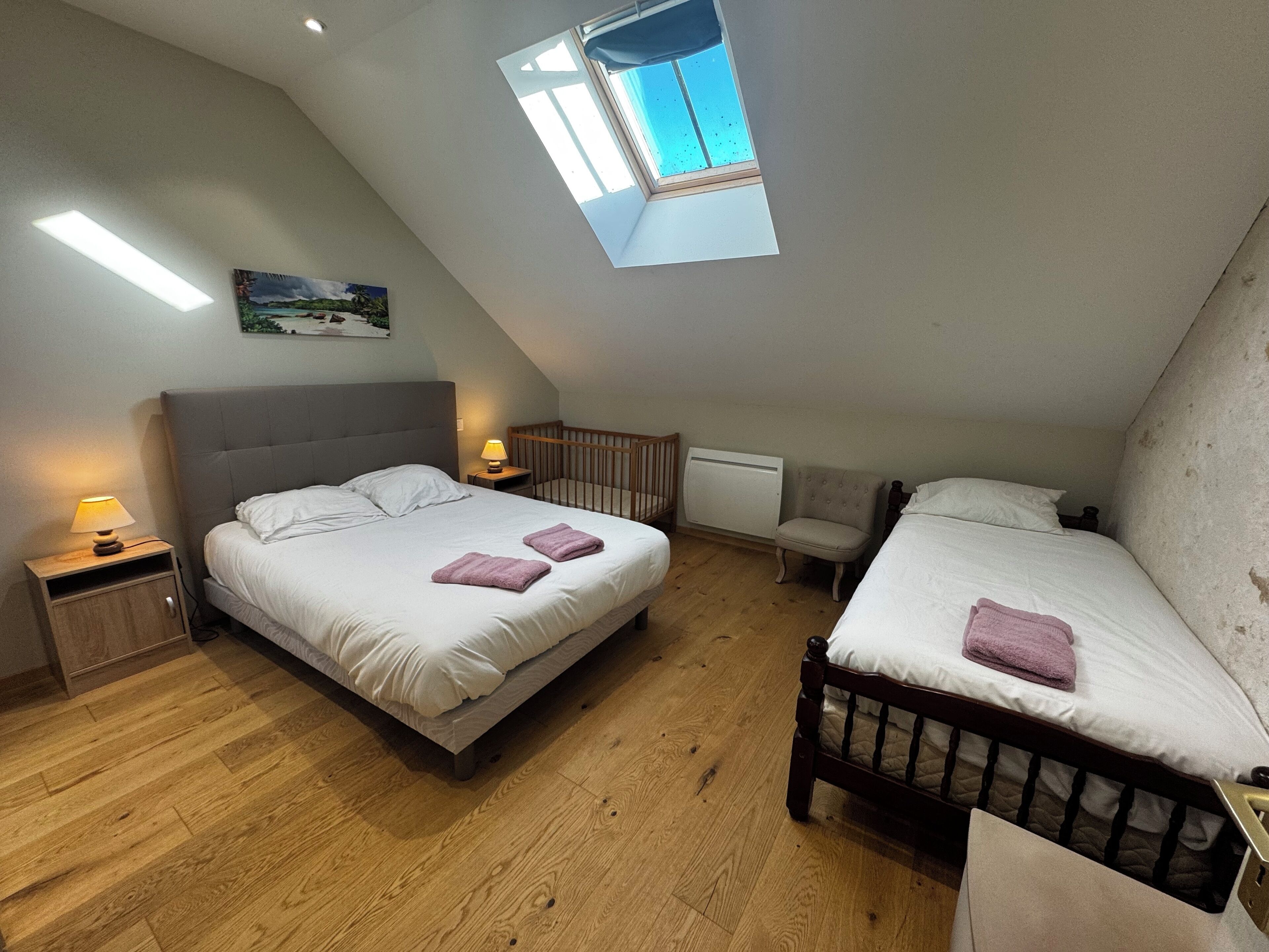 Une chambre avec deux lits, un puits de lumière, une table de chevet avec une lampe, un berceau en bois et un radiateur.