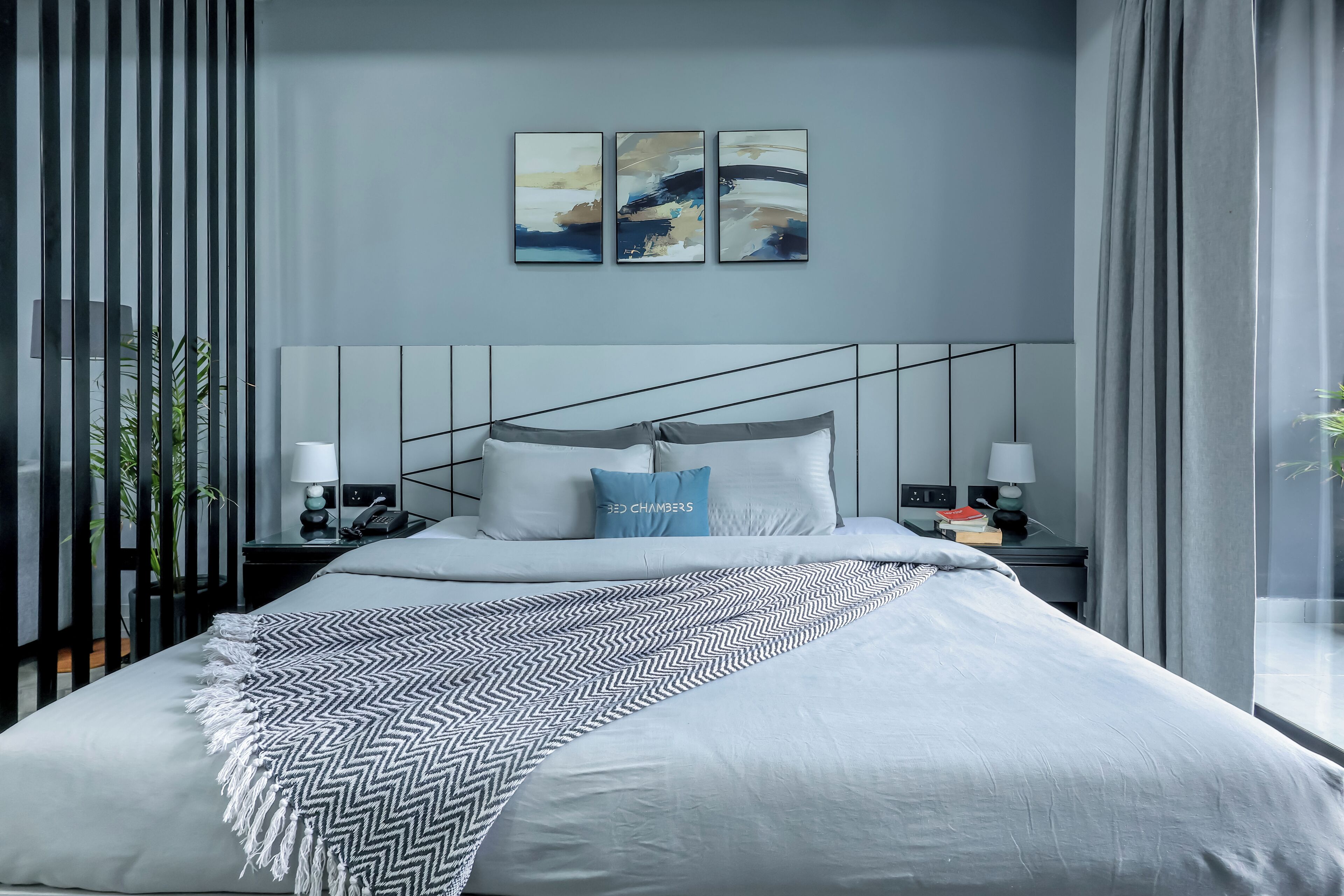 A modern bedroom with a large bed, a headboard with geometric lines, bedside tables with lamps, and abstract wall art.