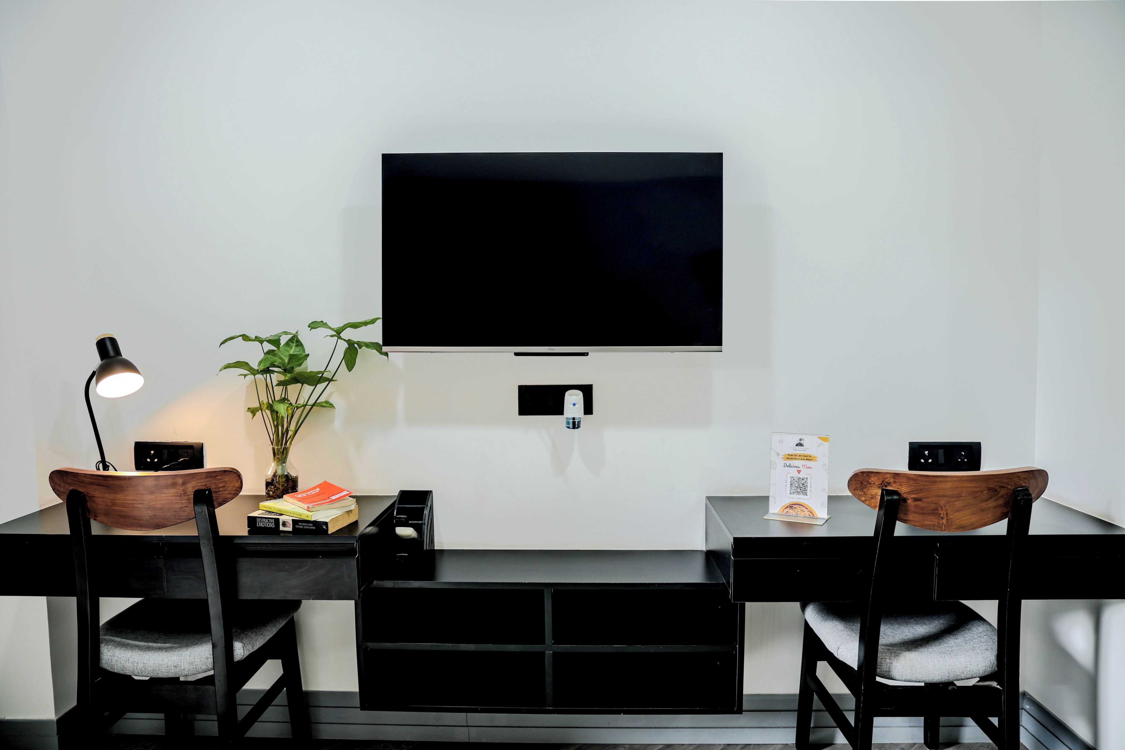 A modern workspace with a flat-screen TV, two wooden chairs, a desk, a lamp, a potted plant, and a small shelf with books and a box.