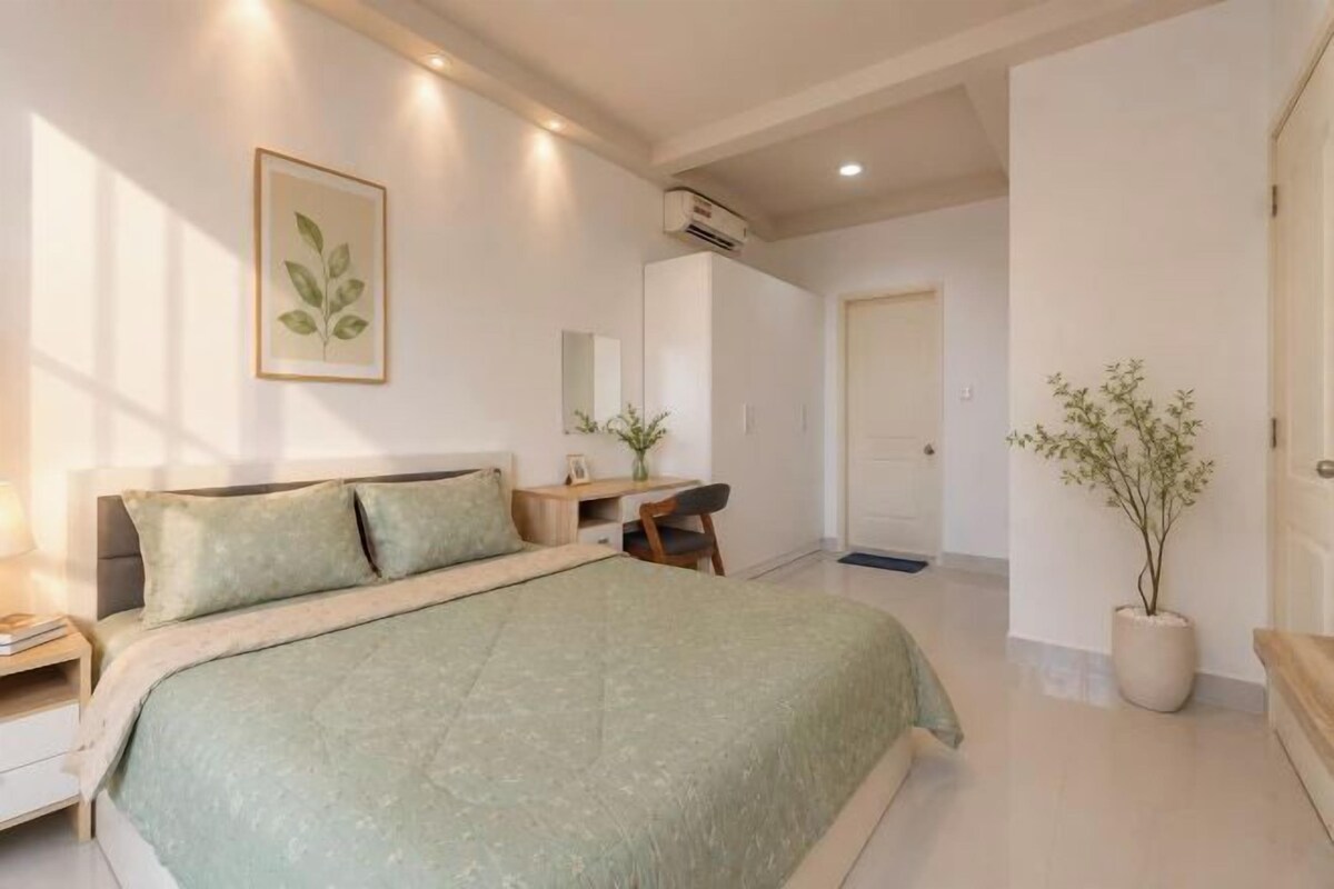 A bedroom with a bed, bedside table, desk, chair, potted plant, and a door.