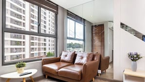 Un salon moderne avec un canapé en cuir marron, une petite table blanche ornée d’une plante et une grande fenêtre donnant sur un immeuble.