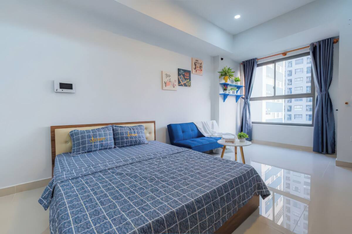 Une chambre moderne avec un grand lit, un fauteuil bleu, une petite table avec une plante et une fenêtre avec des rideaux.