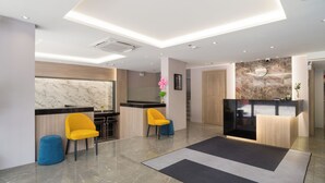 Un hall d’hôtel moderne avec un comptoir d’accueil, deux fauteuils jaunes, un pouf bleu et un sol en marbre.