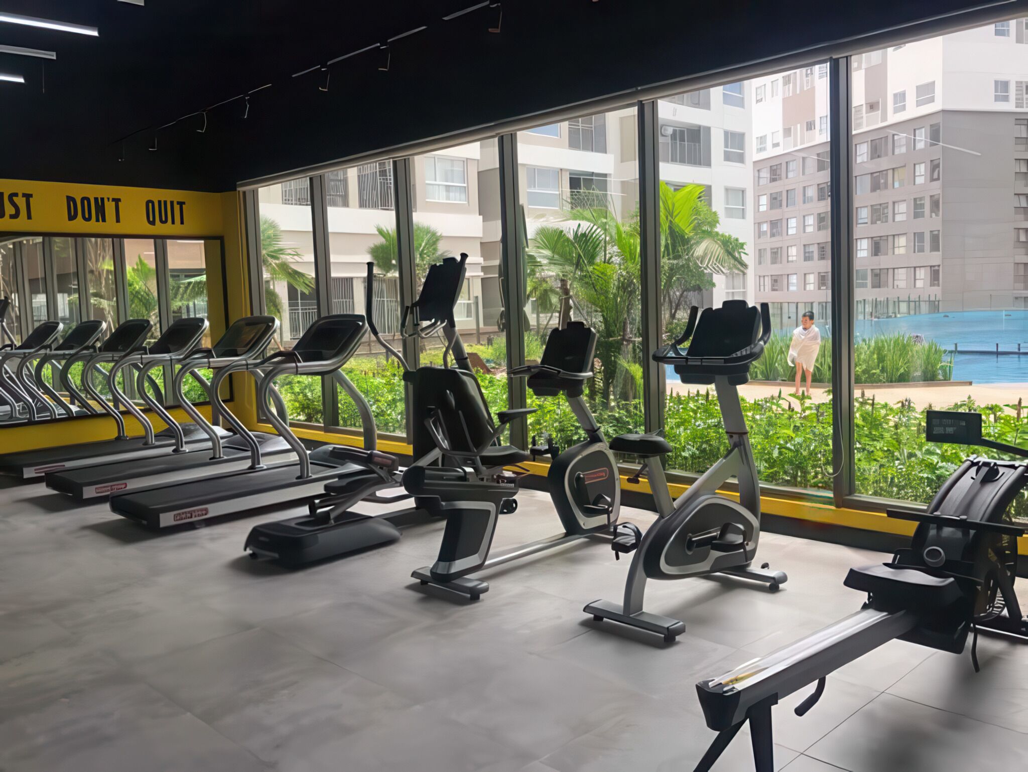 A gym with treadmills and elliptical machines, large windows with a view of a residential building and a swimming pool.