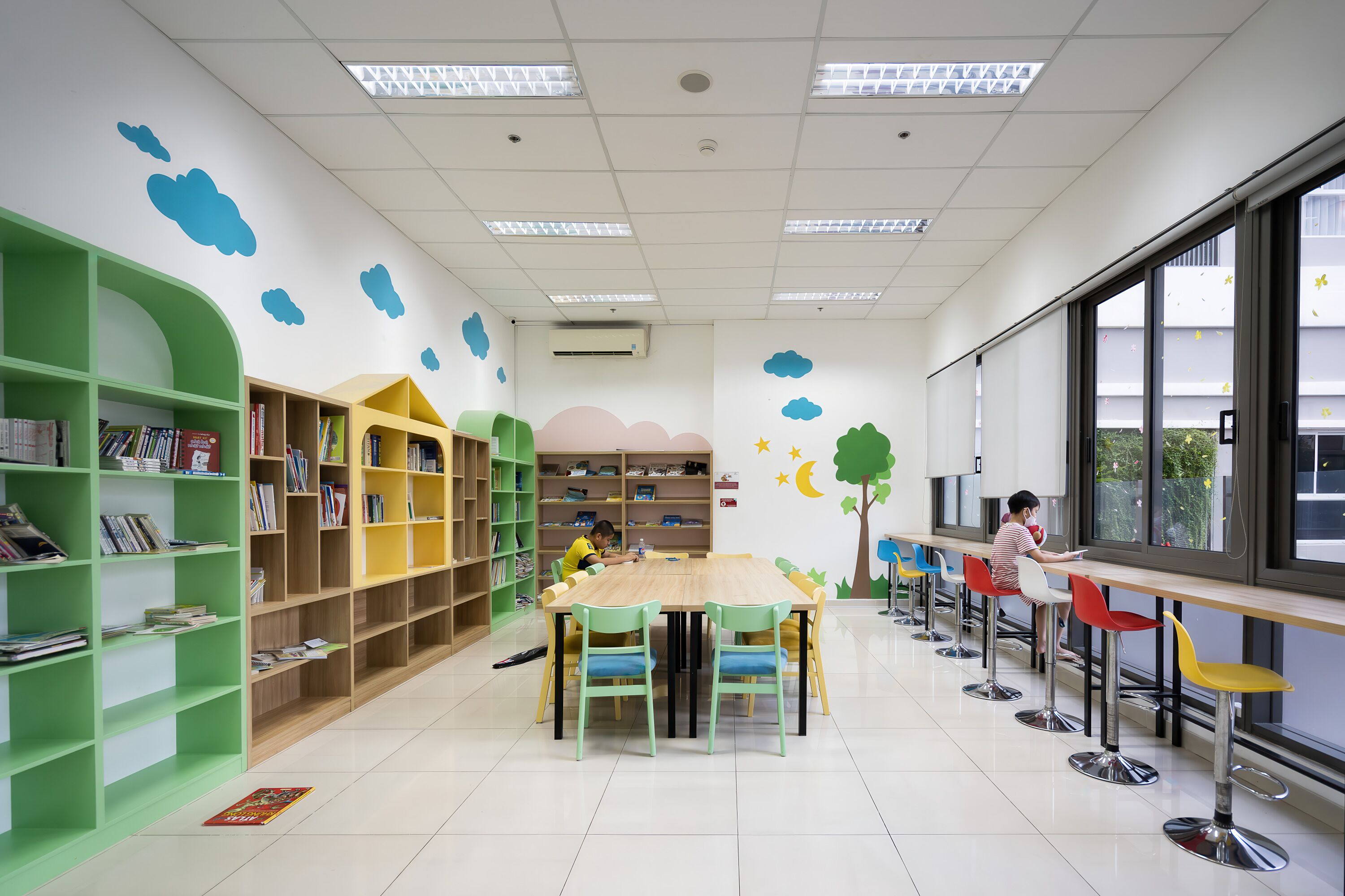 A library with green and yellow bookshelves, a wooden table with chairs, and a large window with a view of the outside.