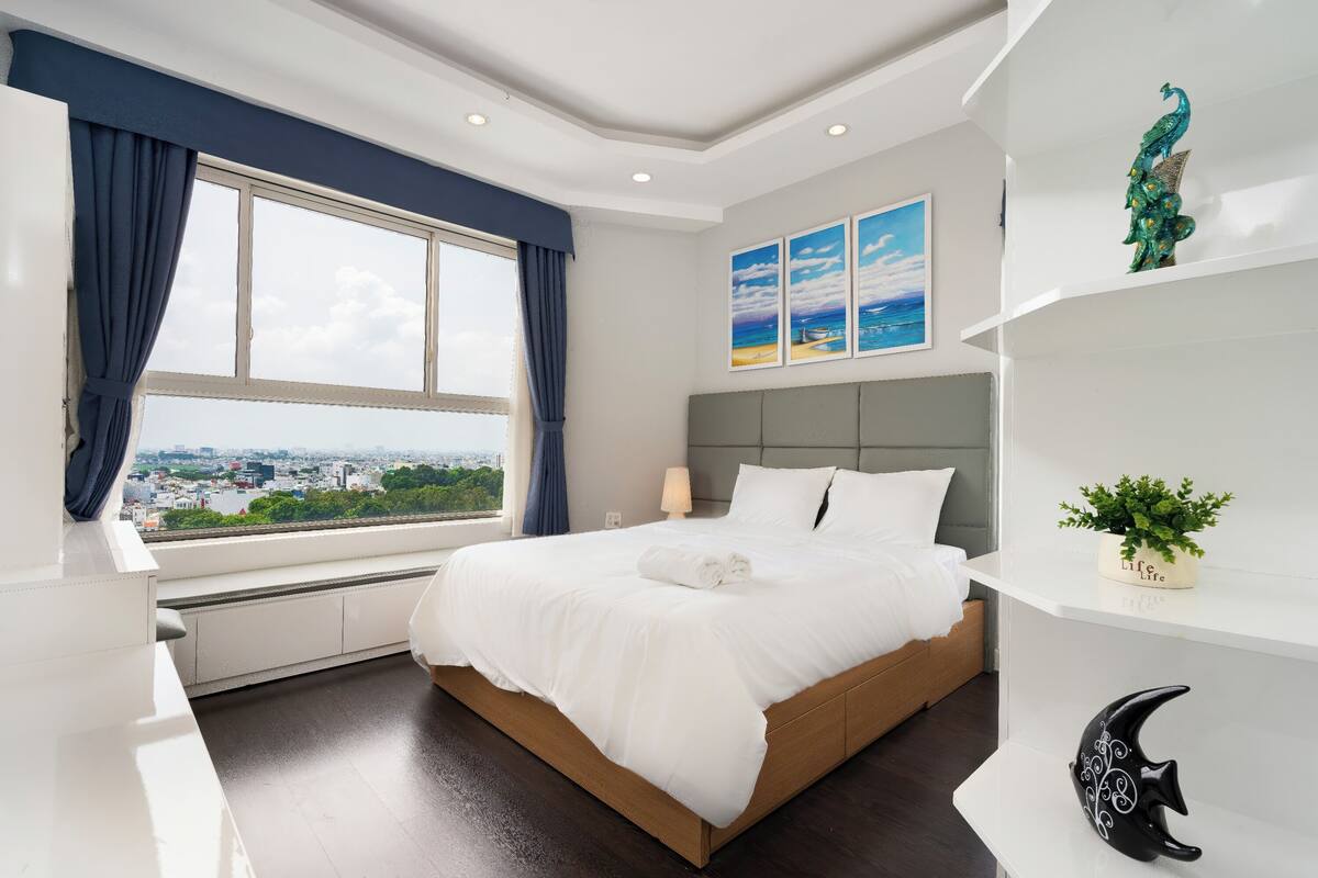 Quarto de hotel moderno com uma cama grande, vista para a cidade e uma estante decorativa com uma planta e uma escultura.