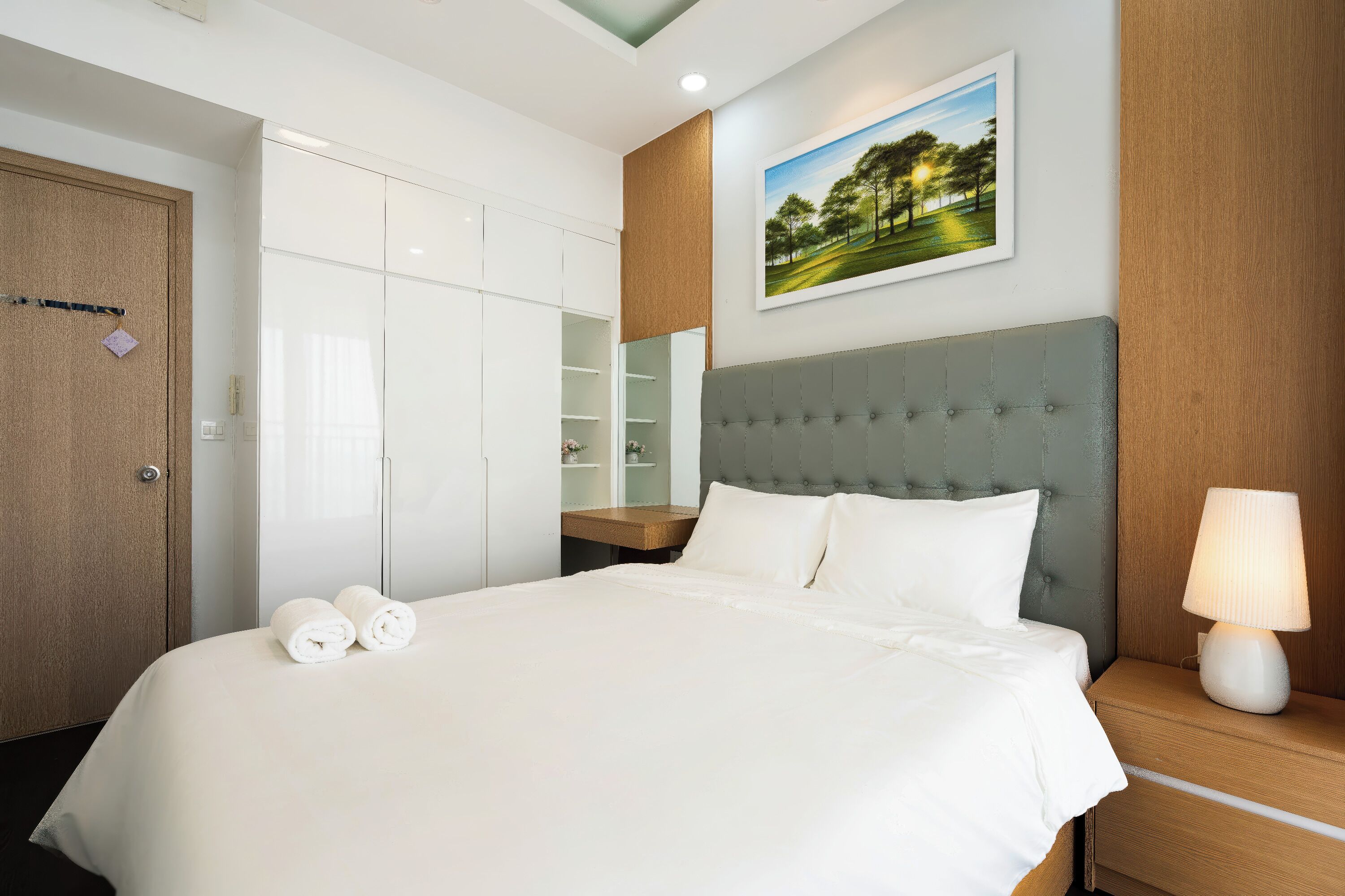 A hotel room with a large bed, a nightstand with a lamp, a wardrobe, a mirror, and a painting on the wall.