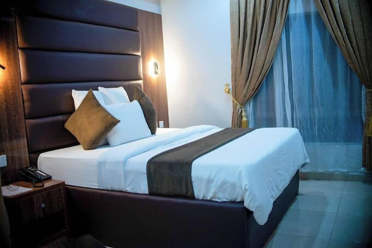 Una habitación de hotel con una cama grande, cabecera de madera, mesita de noche con teléfono y ventana con cortinas.