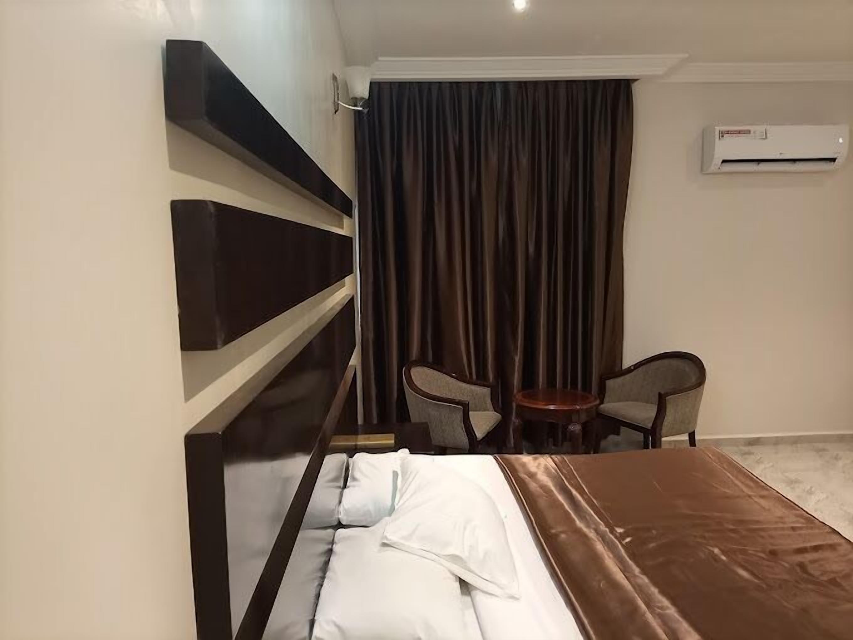 Habitación de hotel con cama, una mesita, dos sillas, cortina y equipo de aire acondicionado.