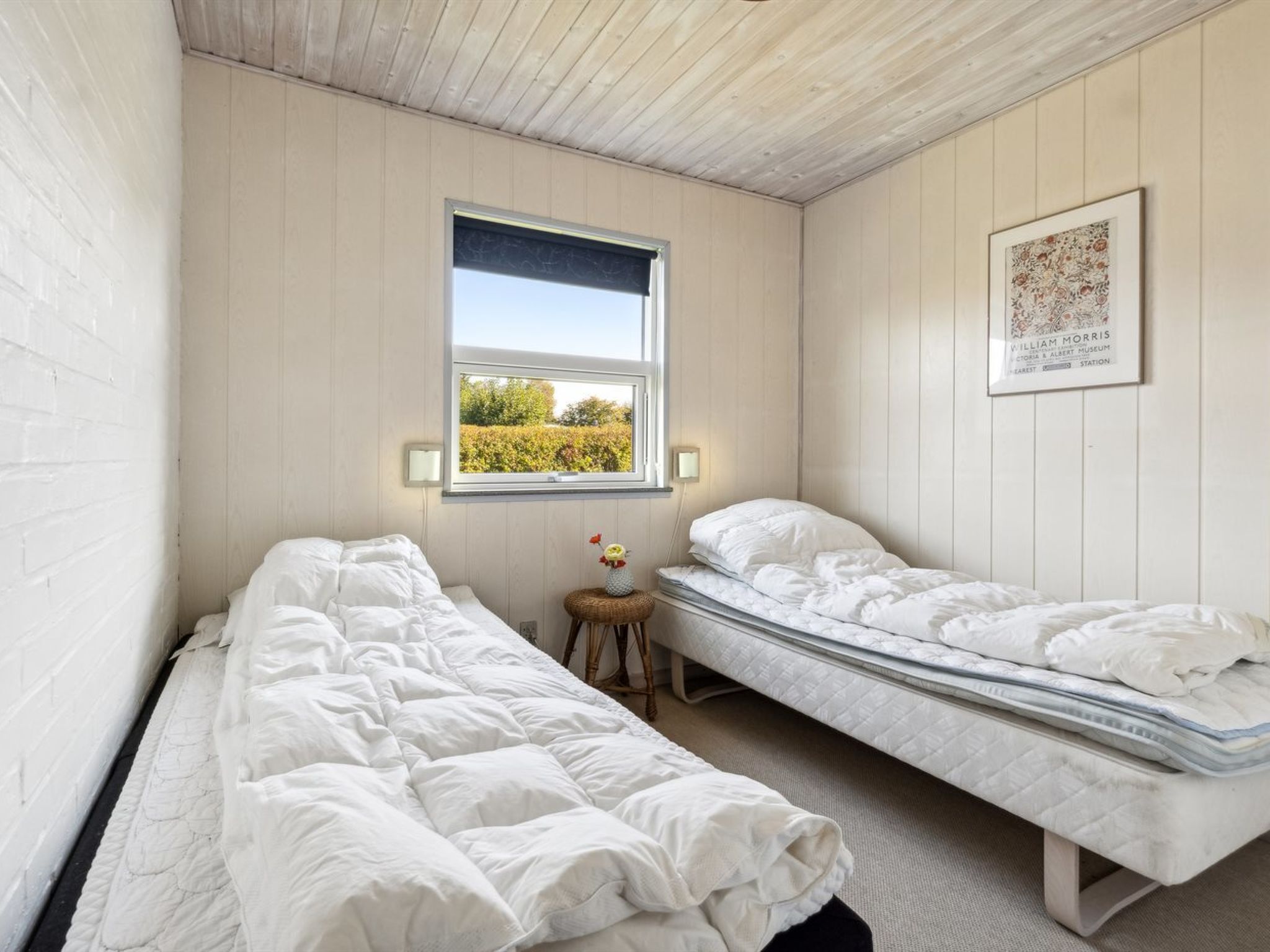 A room with two single beds, a small stool, a window with a view of greenery, and a framed picture on the wall.