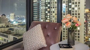 Une chambre moderne dotée d’une grande fenêtre, d’un coin salon confortable, d’une petite table avec un livre et des fleurs, et offrant une vue sur le paysage urbain la nuit.