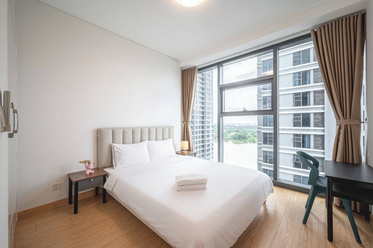 Une chambre d’hôtel dotée d’une grande fenêtre, d’un lit avec du linge de maison blanc, d’une table de chevet avec une lampe et offrant une vue sur le paysage urbain.