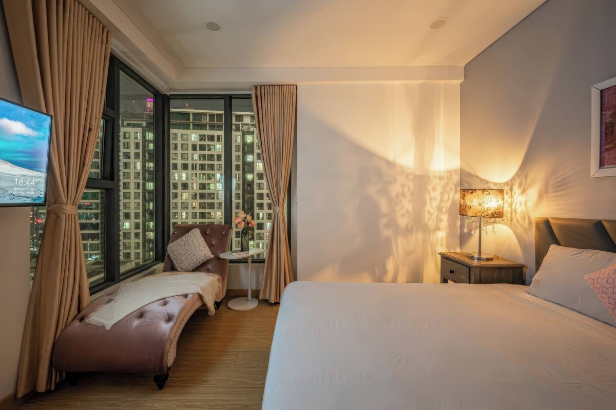Une chambre d’hôtel avec un grand lit, un petit canapé, une table de chevet avec une lampe et une vue sur le paysage urbain à travers de grandes fenêtres.