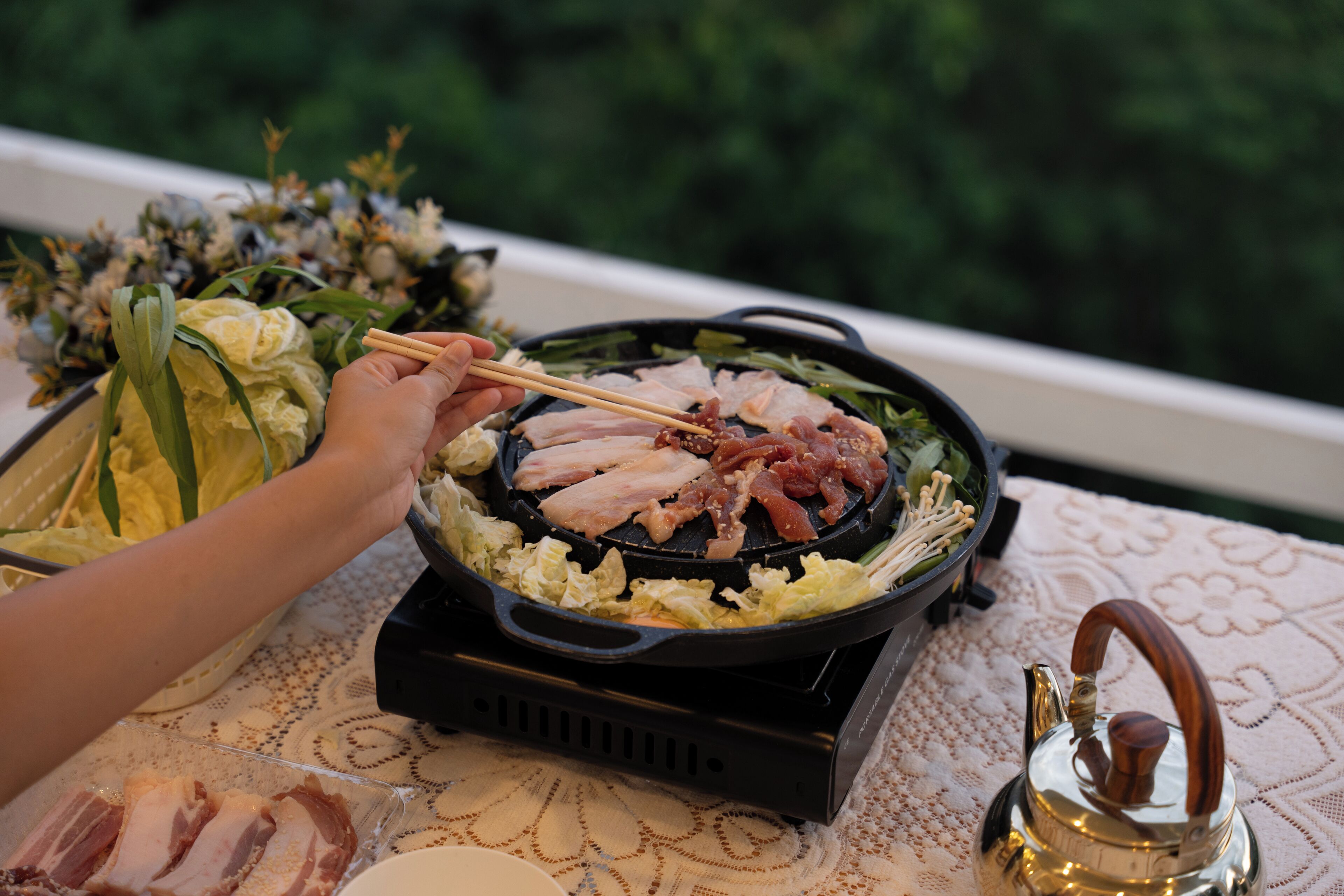 A person grilling meat on a portable stove with a teapot nearby.