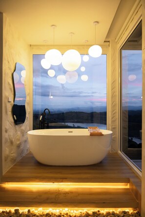 Een moderne badkamer met een vrijstaand bad, een groot raam met uitzicht op een landschap en een bijzondere spiegel.