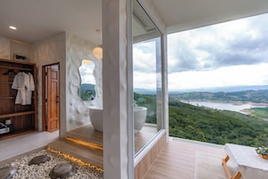 Un baño moderno con una bañera grande, una estantería de madera con toallas y vistas a un río y montañas a través de un ventanal amplio.