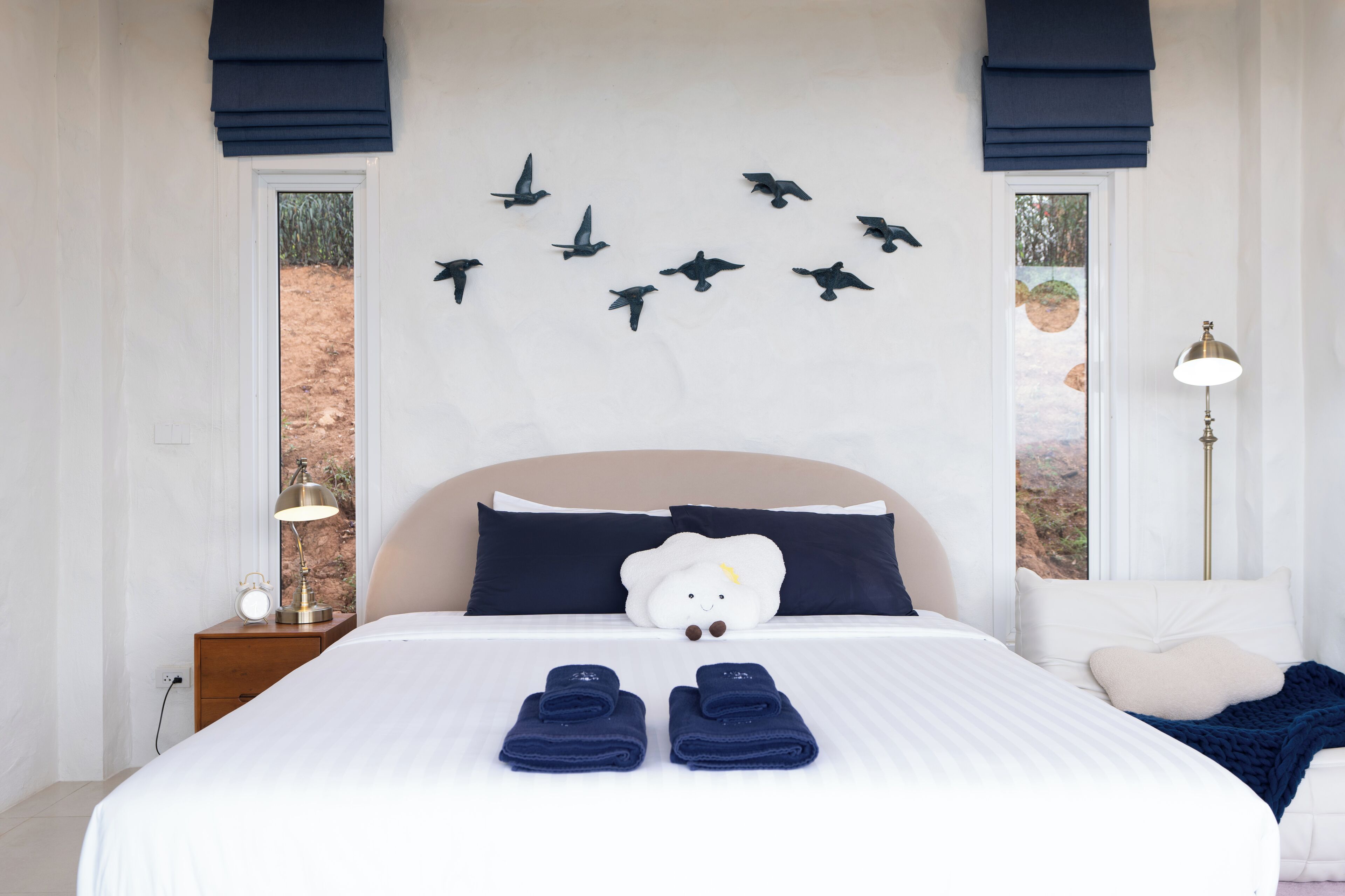 Un lit bien fait, avec une couette blanche et deux serviettes pliées, une table de chevet avec une lampe et une décoration murale représentant des oiseaux.