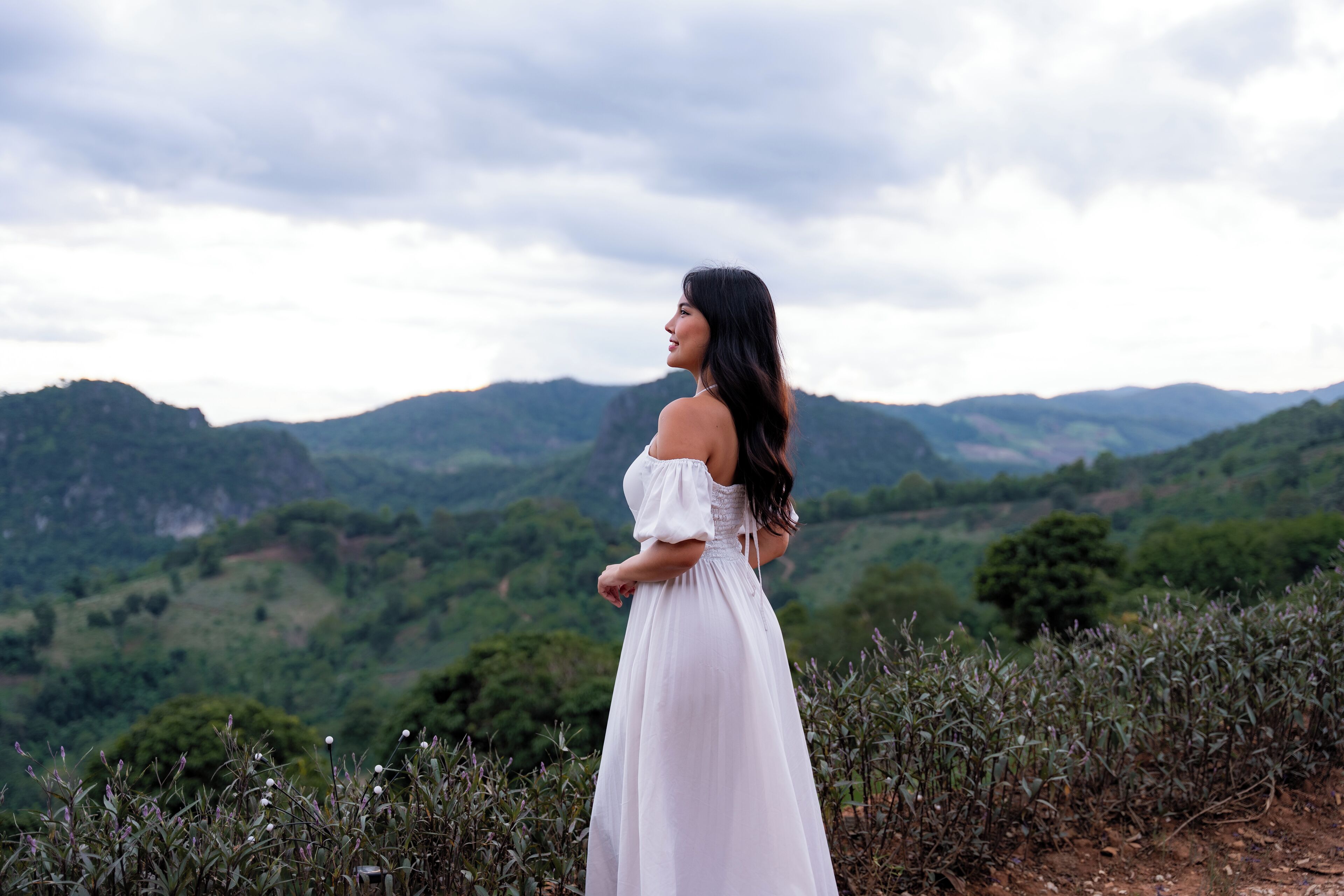 Una persona con un vestido blanco parada en un sendero, con un paisaje montañoso al fondo.