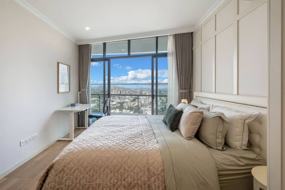 A modern bedroom with a large bed, a desk, and a view of a cityscape through large windows.