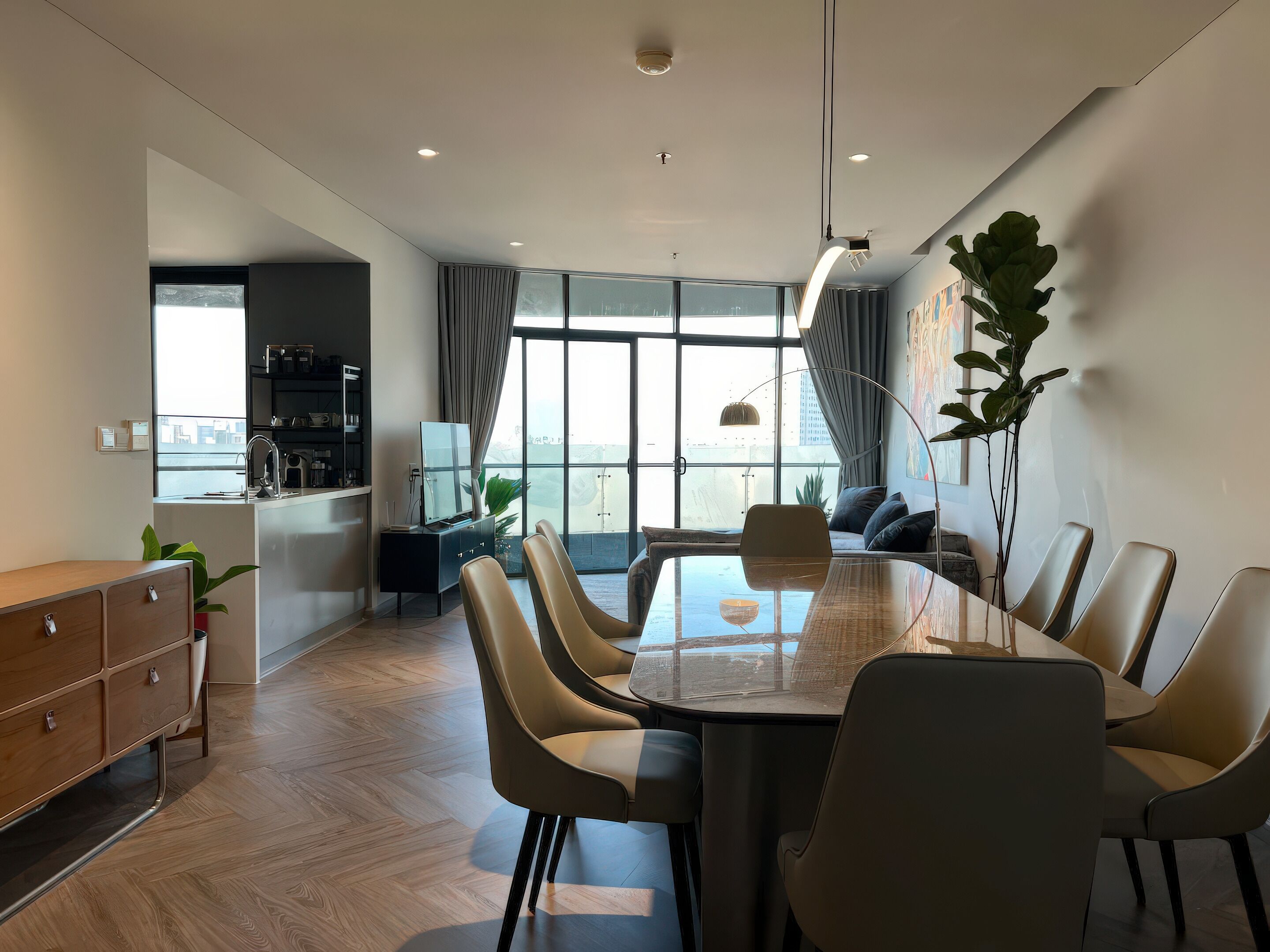 A modern, well-lit dining area with a glass table and comfortable chairs, a kitchen with a built-in fridge and shelves, and large windows offering a view of the city.