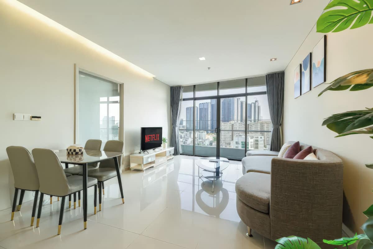A modern living room with a dining area, a flat-screen TV, and a large window offering a city view.