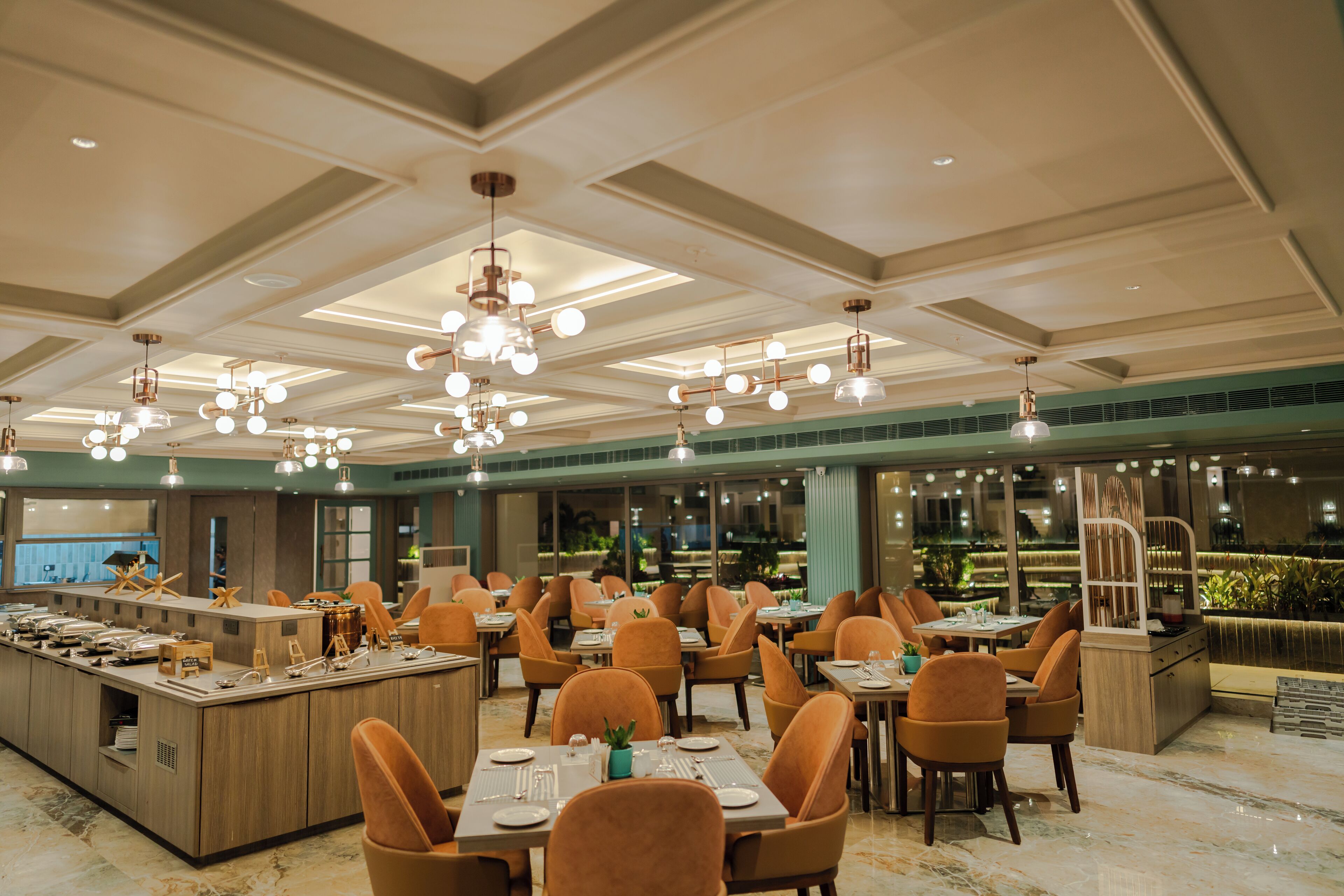 Un restaurant spacieux, doté de nombreuses tables et chaises, d’un îlot central où sont disposés les plats et les boissons, et d’un plafond moderne agrémenté de luminaires suspendus.