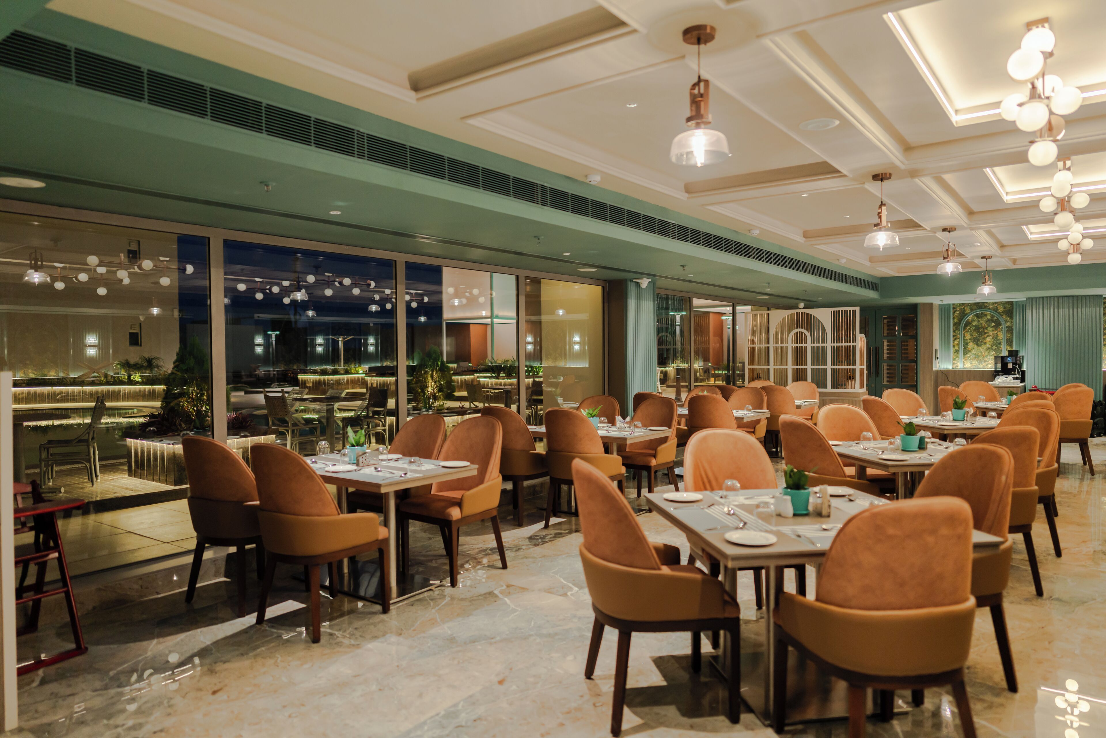 A spacious restaurant with multiple dining tables and chairs, large windows offering views of the night sky, and a modern ceiling design.