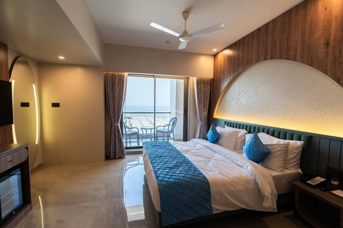 Een moderne hotelkamer met een groot bed, een plafondventilator, uitzicht op zee vanaf het balkon, en een houten accentmuur.