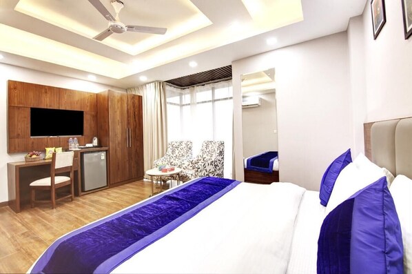 A hotel room with a bed, a desk, a chair, a small table, a TV, and a ceiling fan. - The Majesty Hotel (New Delhi)