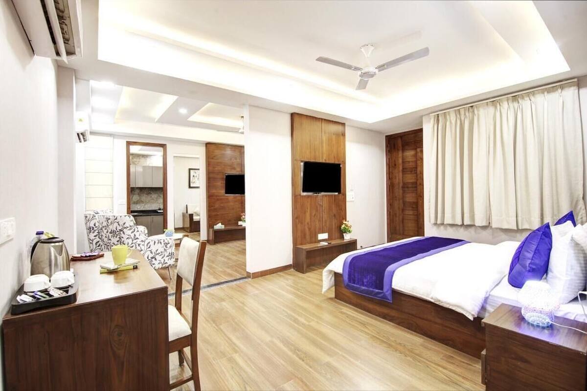 A modern hotel room with a wooden floor, a bed with a blue bedspread, a desk with a kettle, and a television mounted on the wall.