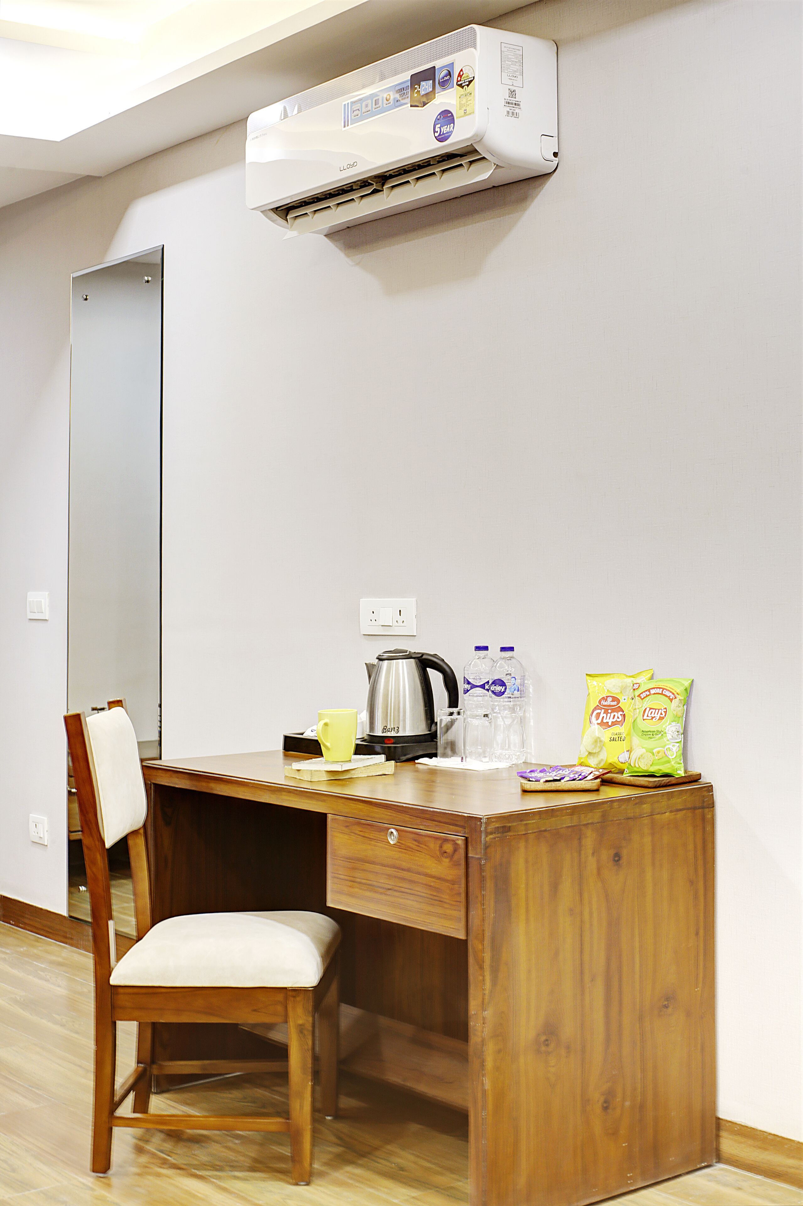 A small wooden desk with a chair, a kettle, a mug, two bottles of water, and snack packets.
