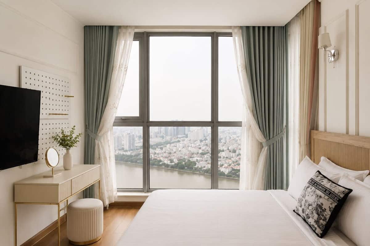 A hotel room with a large window, a bed, a bedside table with a mirror, a TV, and a wall-mounted light fixture.