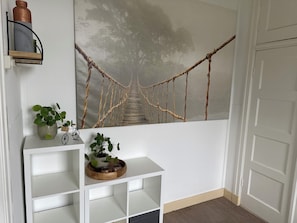 A room with a white wall, a shelf with plants, a small bicycle, and a rope bridge painting.