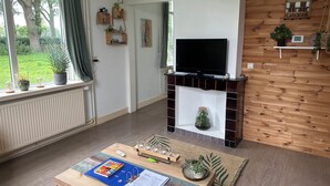 Ein Wohnzimmer mit Kamin, Fernseher, Couchtisch mit Buch und Planer sowie Blick auf Grünfläche.