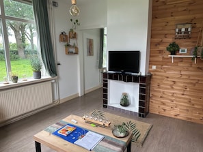 A living room with a fireplace, a television, a coffee table with a book and a planner, and a view of greenery outside.