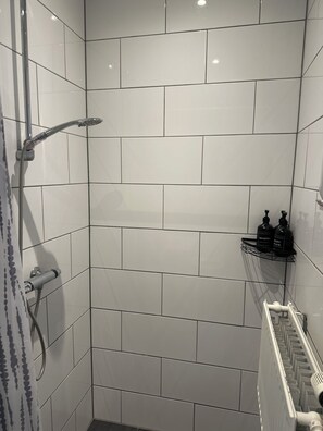 Suihkussa on käsisuihkupää, seinään kiinnitetty hana ja pyyheteline, jossa on myös pulloja.