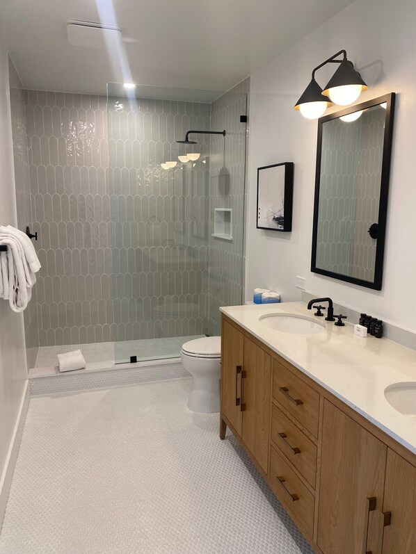 Un baño moderno con cabina de ducha de vidrio, inodoro blanco y tocador de madera con dos lavamanos.