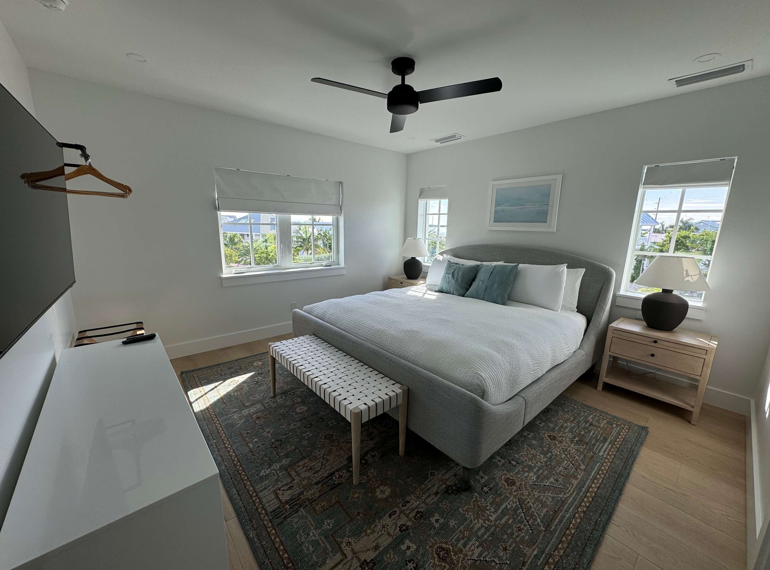 Un dormitorio con cama, mesitas de noche, un banco, ventilador de techo y ventanas con vistas.