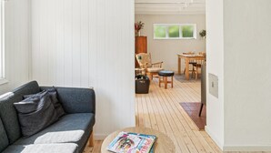 Una sala de estar moderna con un sofá gris, una mesa de centro de madera con una revista, y un comedor con mesa y sillas de madera.